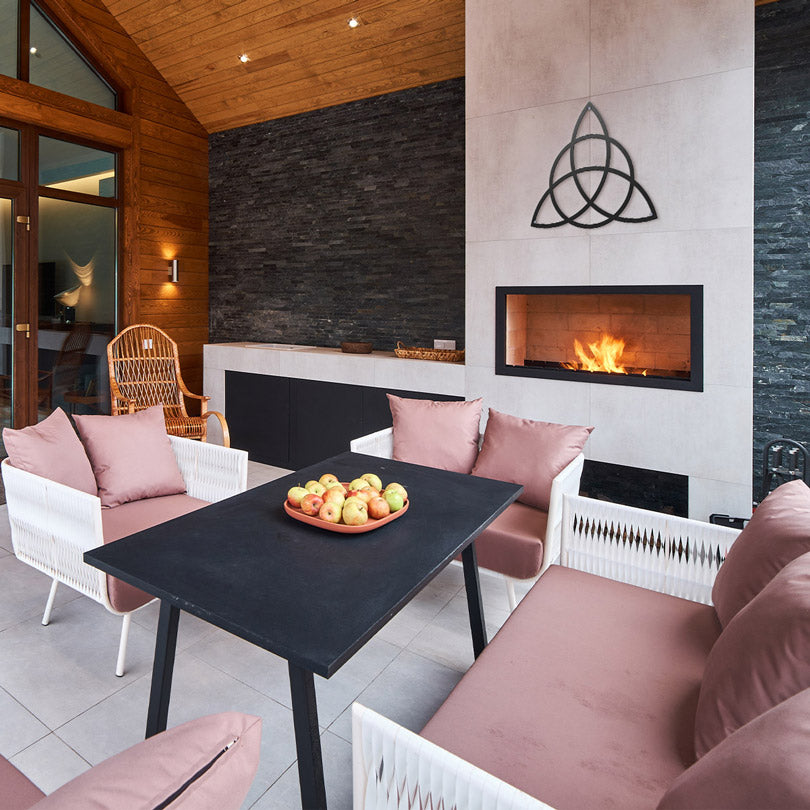 Home Decor Art Wall Sacred Geometry Triquetra Wall Metal Art in black, is displayed In the living room, below is a fireplace, with a burning fire, a sitting area with pink sofas, a black table with a plate of apples.