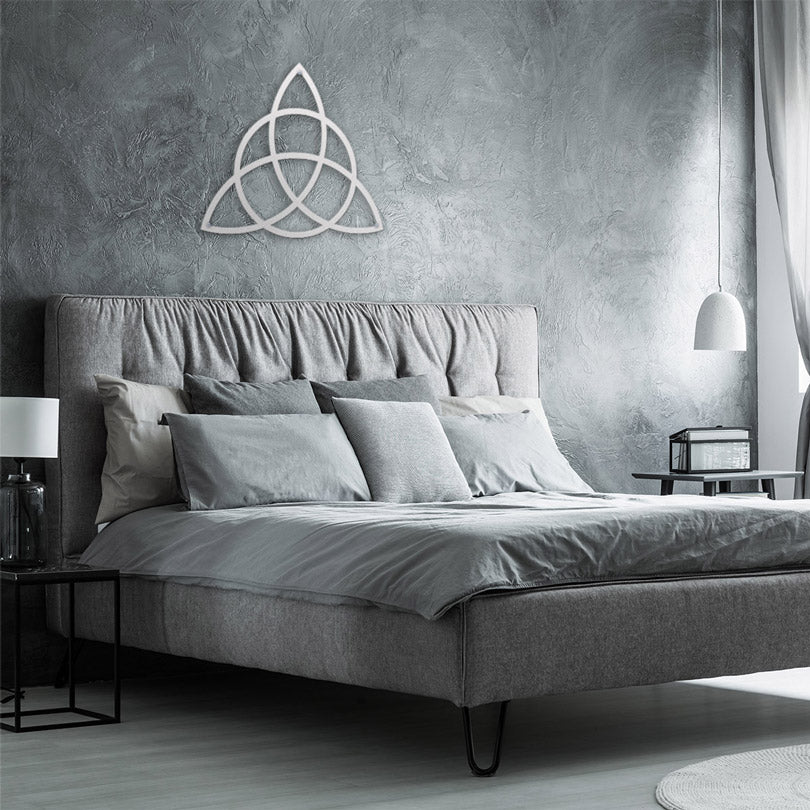 Home Decor Art Wall Sacred Geometry Triquetra Wall Metal Art in white, is displayed in a fashionable bedroom, on a gray wall, with a gray bed, gray decorative pillows, a window with natural light and gray curtains.
