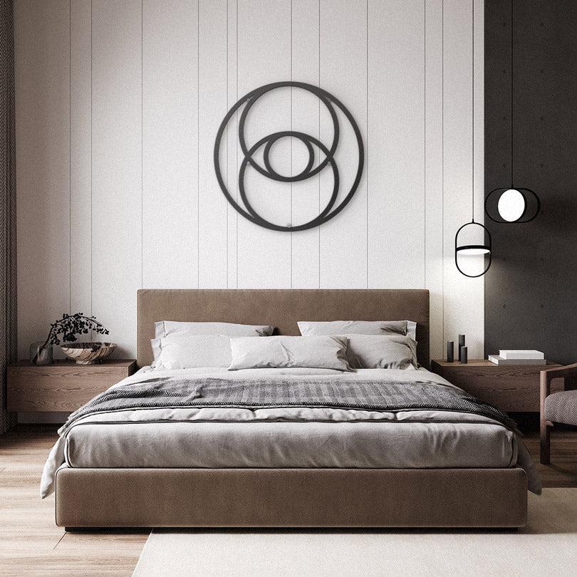 Sacred Geometry Home Decor The Vesica Pisces Metal Wall Art in black is hanging in a modern bedroom with a bed with gray bedding, two black lamps hanging from the ceiling, and two wooden dressers by either side of the bed.